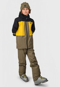 Mustast ja kollasest suusajope esiosal lukuga, koos oliivroheliste suusapükstega. Jopel on logo ja hallid suusasaapad.