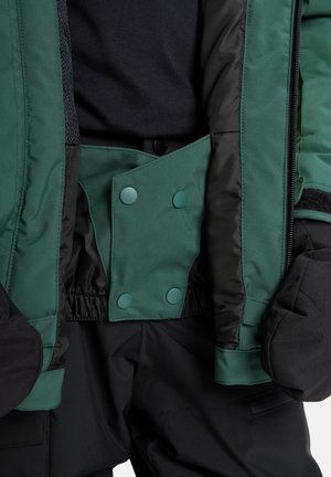 Grüne isolierte Jacke mit schwarzer Innenfütterung, ausgestattet mit Druckknöpfen und elastischen Bündchen, kombiniert mit schwarzer Hose und Fäustlingen.
