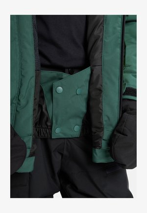 Grøn isoleret jakke med sort foring, med trykknapper og elastiske manchetter, kombineret med sorte bukser og luffer.