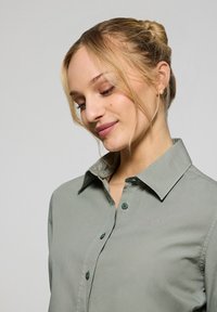 Ljusgrön skjorta med knappar och krage, mjuk textur och subtil logodetalj; har matchande gröna knappar.