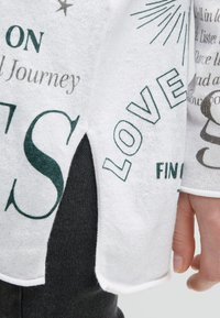 Nahaufnahme eines weißen Stoffärmel mit schwarzem Text und grafischen Drucken, der über einer dunklen Hose getragen wird, wobei eine Person mit der entspannten Hand an ihrer Seite zu sehen ist.