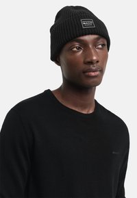 Svart stickad mössa med ribbat mönster och en vit rektangulär logopatch. Svart tröja med rund hals synlig under mössan.