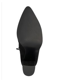 Zapato de tacón alto de ante negro con punta afilada y suela texturizada. Presenta una silueta elegante y una almohadilla en el talón redondeada para mayor agarre.