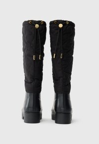 Bottes noires matelassées hautes avec cordons ajustables et détails en métal doré, vues de l'arrière sur un fond uni.