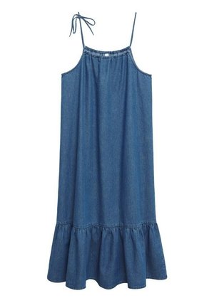 Kvinde iført en ærmeløs blå denim midi-kjole med flæsekant og beige strop-sandaler, stående mod en ensfarvet hvid baggrund.