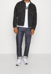 Veste en denim noire à motifs, t-shirt blanc avec logo, pantalon en denim foncé, et baskets blanches avec des accents teal. Tenue décontractée dans l'ensemble.