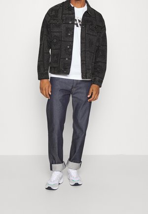 Veste en denim noire à motifs, t-shirt blanc avec logo, pantalon en denim foncé, et baskets blanches avec des accents teal. Tenue décontractée dans l'ensemble.