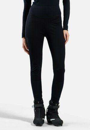 Person wearing black fitted leggings and black ski boots standing on a white background.