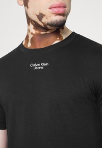 T-shirt en coton noir à manches courtes et col rond. Présente un logo blanc "Calvin Klein Jeans" sur la poitrine. Texture lisse.