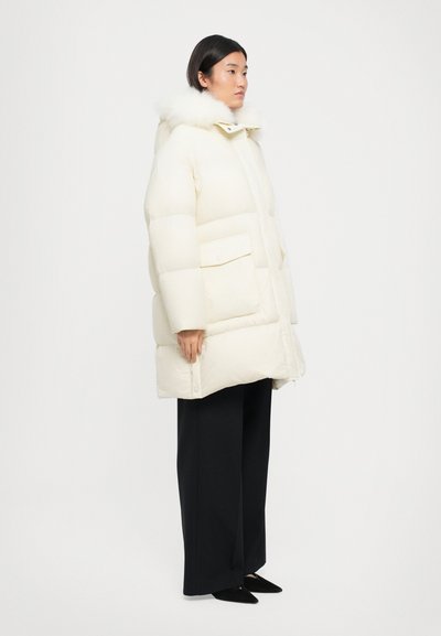 Weiße, plüschige Jacke mit einer mit Fell gefütterten Kapuze, locker geschnitten, mit großen Fronttaschen und einer glatten, glänzenden Oberfläche. Schwarze Hosen vervollständigen das Outfit.