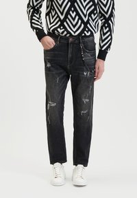 Zwarte versleten denim jeans met een tapered fit, voorzien van rafelige details en een zilveren kettingaccent, gecombineerd met witte sneakers.