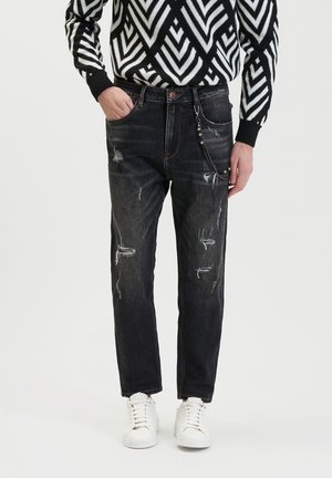 Zwarte versleten denim jeans met een tapered fit, voorzien van rafelige details en een zilveren kettingaccent, gecombineerd met witte sneakers.