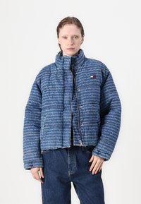 Gequiltede blauwe pufferjas met een logo print over het hele oppervlak, hoge kraag, ritssluiting en zijzakken. Gepaard met donkere spijkerbroek.