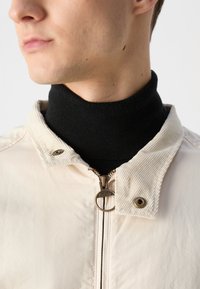 Beige Jacke mit einem Cordkragen, metallischem Reißverschluss und schwarzem Rollkragenpullover darunter. Der Stoff wirkt glatt mit minimalen Texturdetails.