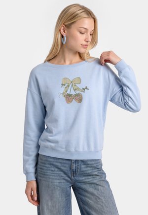 Ung kvinde iført lyseblå sweatshirt med kirsebær- og sløjfegrafik, lyseblå øreringe i cirkel, og blå jeans, kigger ned og rører ved skulderen.