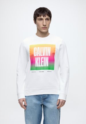Fiatal férfi fehér pulóvert visel színes "Calvin Klein" logóval, világoskék farmernadrággal párosítva, fehér háttér előtt áll.