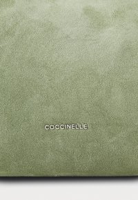 Bolsa em camurça de cor verde clara, com uma textura suave e o nome da marca "Coccinelle" em relevo prateado, posicionado centralmente na parte inferior.