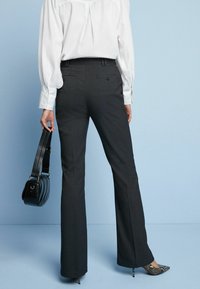 Schwarze, taillierte Hose mit leichtem Schlag, aus einem glatten Stoff. Kombiniert mit einer weißen Bluse und einer kleinen schwarzen Handtasche.
