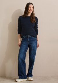 Mörkblå stickad tröja med en strukturerad design, matchad med blå raka jeans och vita sportskor, mot en neutral bakgrund.