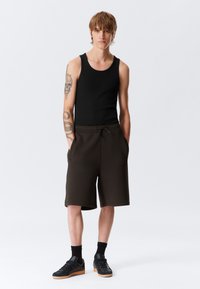 Un maiou negru purtat împreună cu pantaloni scurți maro închis. Pantalonii scurți au un brâu elastic și buzunare laterale. Modelul poartă adidași negri cu tălpi din cauciuc.