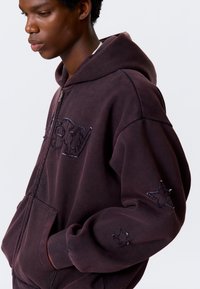 Bordeauxrode hoodie met rits van zachte stof, met geborduurde tekst op de borst en steraccenten op de mouwen.