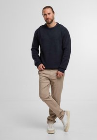 Maglione a maglia navy con scollo rotondo, pantaloni beige a gamba dritta e sneakers bianche con dettagli beigi. Il modello si trova su uno sfondo neutro.
