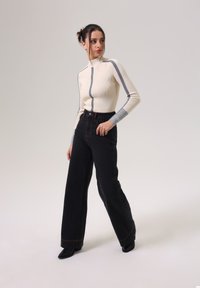 Cremig gerippter Rollkragenpullover mit grauen Akzenten, kombiniert mit hoch-taillierten, schwarzen weit geschnittenen Jeans mit orangefarbenen Nähten und spitzen schwarzen Schuhen.