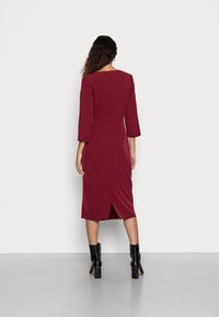 Robe midi bordeaux avec des manches trois-quarts, fente au dos pour plus de liberté de mouvement, et une silhouette ajustée. Associée à des bottines noires.