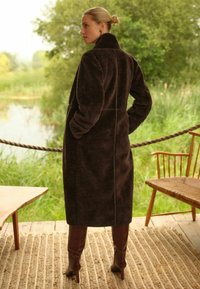 Barna műszőrméből készült kabát magas gallérral, oldalzsebekkel és hosszú, kényelmes szabással. A modellen térdig érő barna csizma van. Természetes háttér.