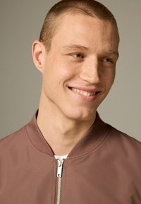 Brązowa kurtka bombowca z zapięciem na zamek, prążkowanym kołnierzem i gładką fakturą. Model ma ogoloną głowę i lekko się uśmiecha.