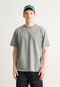 Gray cotton t-shirt with a round neckline and short sleeves, paired with dark pants. The fabric has a soft texture and relaxed fit.