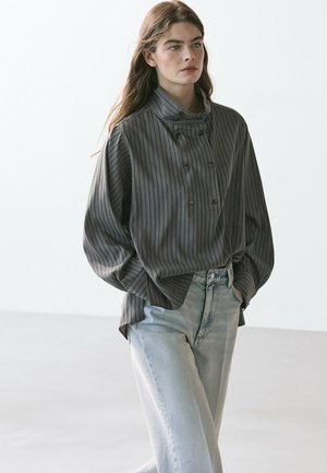 Frau mit langen Haaren, die ein lockeres graues, gestreiftes, geknöpftes Oberteil und hellblaue Jeans trägt, mit den Händen in den Taschen vor einem einfarbigen Hintergrund stehend.