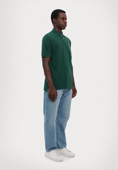 Polo verde de algodón, con collar a rayas y un pequeño diseño bordado en el pecho, combinado con jeans azules y zapatillas blancas.