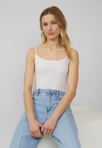 Weißes Tanktop mit dünnen Trägern und rundem Ausschnitt, kombiniert mit hellblauen hochgeschnittenen Jeans. Der Stoff wirkt glatt und eng anliegend.