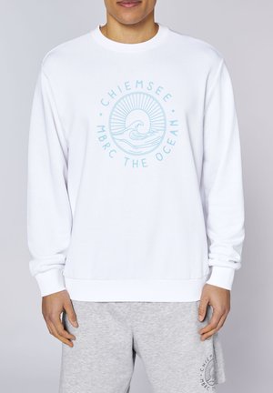 Man wearing white sweatshirt with light blue ocean wave and sun graphic and text "Chiemsee MBRC The Ocean," paired with gray shorts.