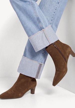 Personne portant un jean bleu clair avec revers et des bottines en daim marron avec fermetures éclair latérales et talons bas, debout sur un sol blanc.