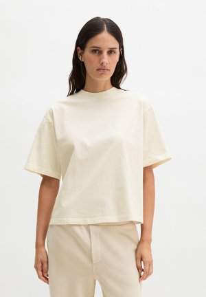 Kobieta o długich ciemnych włosach, ubrana w luźny, beżowy t-shirt z krótkim rękawem i jasnobeżowe spodnie, stojąca na tle gładkiej białej ściany.