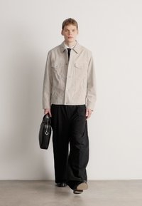 Világosszürke velúr dzseki két mellzsebbel, laza fekete nadrággal párosítva. A modell egy fekete bőr táskát tart a kezében. Semleges háttér.