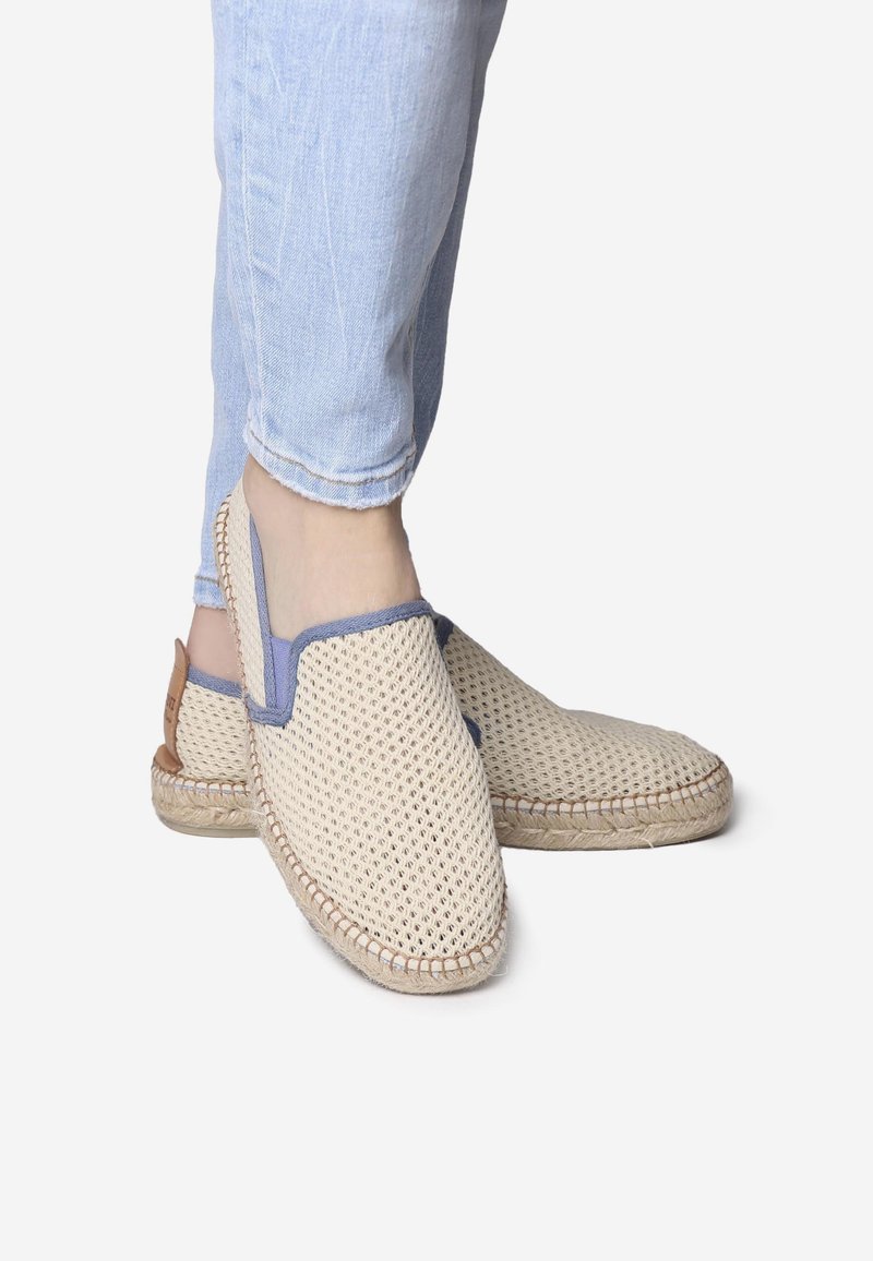 Osoba oblečená v světle modrých džínách a béžových tkaných slip-on botách s modrým lemováním, stojící proti bílé pozadí.