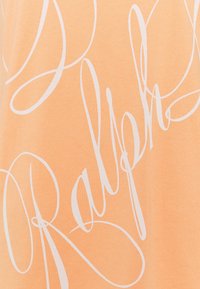 Ljusorange t-shirt i mjukt tyg med stora vita skrivstilsbokstäver som säger "Ralph" med flödande, sammanlänkade svängningar.