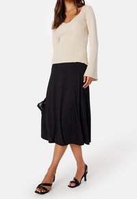 Beige ribbattu pitkähihaine paita V-kaula-aukolla ja taskulla, yhdistettynä laskostettuun mustaan midi-hameeseen. Mustat remmihyppysandalit.