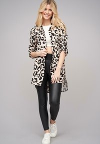 Leopardenmuster langes Hemd in Beige und Schwarz, kombiniert mit schwarzen Leggings und weißen Sneakers. Das Material sieht glatt aus und hat eine lockere Passform.