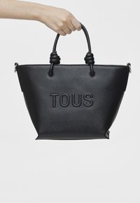 Borsa tote nera in tessuto con manici arrotolati, logo "TOUS" in rilievo sul fronte e forma strutturata. Presenta un attacco laterale per accessori.
