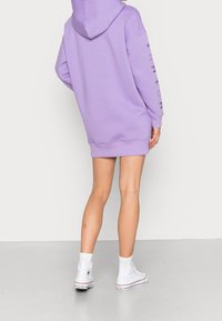 Lila oversized hoodie med snortræk i hætten, ribbede manchetter og kant. Har trykte detaljer på ærmerne og bæres med hvide højhælede sneakers.