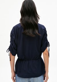 Donna con lunghi capelli scuri, vestita con una maglietta blu navy a maniche corte arricciate e jeans azzurri, in piedi su uno sfondo bianco.