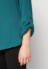 Blouse turquoise à manches longues avec poignets retroussés maintenus par un bouton, en tissu texturé, associée à un pantalon noir.