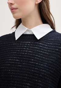 Pull en maille bleu marine avec un tissage ouvert texturé, comportant un col chemise blanc contrastant en dessous.