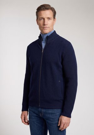 Mann trägt einen marineblauen Pullover mit Reißverschluss, gerippten Ärmeln und Kragen über einem hellblauen Rollkragenpullover und steht vor einem einfarbigen hellen Hintergrund.