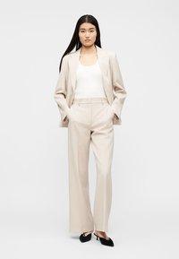 Femeie cu păr lung și negru, purtând un costum bej cu pantaloni largi, top alb fără mâneci și pantofi negri ascuțiți, stând cu mâinile în buzunare.