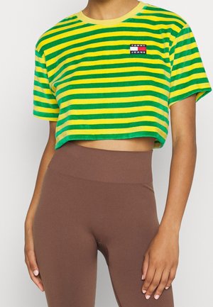 Osoba nosząca zielono-żółtą krótka koszulkę z logo Tommy Jeans i brązowe legginsy z wysokim stanem na białym tle.